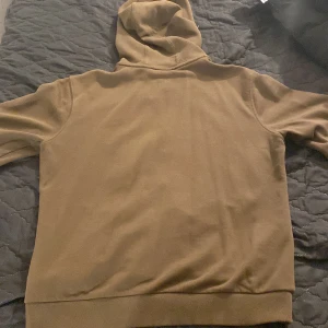 Olivgrön hoodie från Hugo Boss - Säljer en olivgrön hoodie från Hugo Boss med dragkedja framtill och huva med snörning. Tröjan har två fickor och en liten logotyp på bröstet med röd ram. Perfekt för en avslappnad stil.