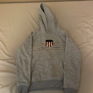 Grå hoodie från GANT/158/164 - Säljer en klassisk grå hoodie från GANT med broderad logga och sköld på bröstet. Tröjan har huva och magficka, perfekt för en avslappnad stil. Mjuk och bekväm med sportig känsla.
