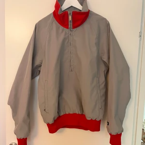 Vintage grå och röd vindjacka från The North Face - Säljer en grå vindjacka från The North Face med röda detaljer på krage, muddar och nederkant. Jackan har halv dragkedja framtill och två sidofickor. Perfekt för blåsiga dagar och har en sportig look.