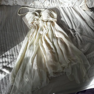 Beige axelbandslös klänning med flätad detalj - Säljer en krämvit axelbandslös klänning med vackra draperingar och flätad detalj längs bysten och som axelband. Klänningen har en luftig, lätt utsvängd kjol och är tillverkad i ett tunt, flowy material som ger en romantisk känsla.