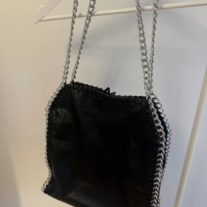 Svart axelväska med kedjerem - Säljer en svart axelväska med silverfärgade kedjeremmar. Tyvärr har det gått upp lite högst upp på väskan men jag är säker på att det går att lösa med lite nål och tråd! Säljs på grund av att den inte används längre❤️
