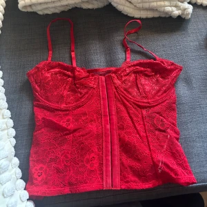 Röd spetskorsett med smala axelband - Säljer en röd spetskorsett med smala justerbara axelband och dekorativ spets över hela plagget. Framsidan har en markerad söm och kupor med lätt stöd. Perfekt för att lyfta en outfit med lite extra edge. Storlek 80B men skulle säga att den passar S, kuporna är något mindre än vanligt 