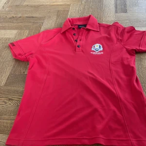 Röd pikétröja Ryder Cup Gleneagles - Röd pikétröja från Ryder Cup 2014 Gleneagles-brodyr på bröstet. Klassisk krage och knappar framtill. Perfekt för golf eller sportiga tillfällen. Bra skick utan typ under armhålan men tog bild på det!