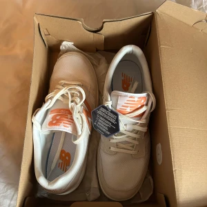 Vita New Balance  - Säljer ett par helt oanvända vita New Balance sneakers med orange logga och detaljer. Skorna har snörning, rund tå och platt sula. Perfekta för dig som gillar stilrena och sportiga skor med en färgklick.