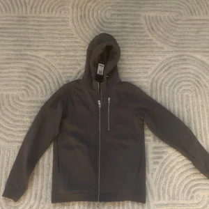 Grå zip Sail Racing - Mörkgrå zip från Sail Racing med dragkedja framtill och huva. Zipen  har en bröstficka med dragkedja och två sidofickor. Perfekt för en avslappnad stil och enkel att matcha med det mesta.