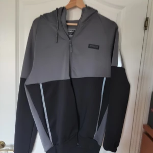 Grå och svart hoodie från McKenzie - Säljer en snygg hoodie från McKenzie i grått och svart med dragkedja framtill och huva. Tröjan har långa ärmar, svarta paneler och reflekterande detaljer på axlarna.