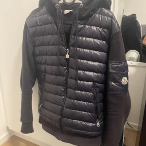 Mörkblå hybridjacka med huva från Moncler - Säljer en mörk blå Moncler-jacka med quiltad front och ärmar i sweatshirtmaterial. Jackan har huva, dragkedja och Moncler-logga på ärmen. Perfekt för dig som vill ha en stilren look.