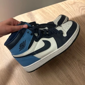 Nike Air Jordan 1 High blå och vita sneakers - Säljer ett par Nike Air Jordan 1 High sneakers i blått, vitt och mörkblått. Klassisk hög modell med snörning och ikonisk swoosh-logga på sidan. Perfekta för dig som gillar streetwear och vill sticka ut med snygga färgblock.