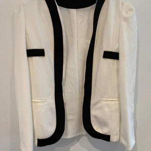 Vit kavaj med svarta detaljer - Snygg vit kavaj med svarta kontrastkanter och dekorativa fickor framtill. Kavajen har lång ärm och en klassisk, stilren design som passar perfekt till en dressad look. Materialet känns strukturerat och ger en exklusiv känsla.