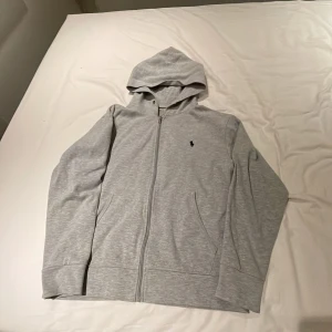 Ljusgrå hoodie från Polo Ralph Lauren - Säljer en ljusgrå hoodie från Polo Ralph Lauren med dragkedja och huva. Tröjan har två fickor framtill och en liten broderad logga på bröstet. Perfekt för en avslappnad stil.