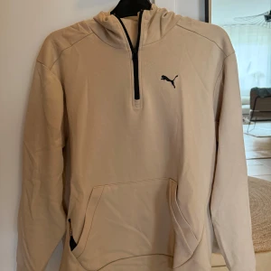 Beige hoodie från Puma - Säljer en beige hoodie från Puma med halv dragkedja framtill och svart Puma-logga på bröstet. Tröjan har huva och en stor magficka. Perfekt skick - inga skador eller liknande.