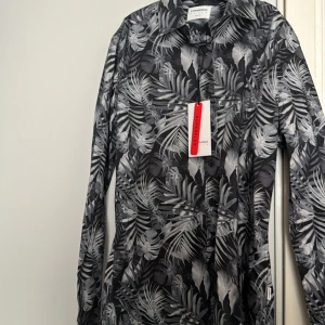 Svart och vit bladmönstrad skjorta från Lindbergh - Säljer en slim fit skjorta från Lindbergh med svart och vit tropiskt bladmönster. Skjortan har långa ärmar och knappar framtill. Perfekt för dig som vill sticka ut med en snygg och modern look.