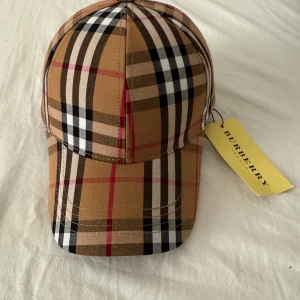 Rutig keps från Burberry - Säljer en klassisk rutig keps från Burberry i beige, svart, röd och vit. Kepsen har den ikoniska Burberry-rutan och justerbar rem baktill. Perfekt accessoar för en trendig look.