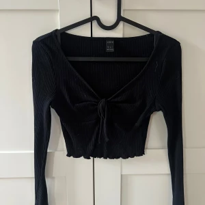 Svart croppad ribbad topp från SHEIN - Svart långärmad croppad topp från SHEIN med ribbad struktur och knyt framtill. Djup v-ringning och vågig nederkant ger en trendig look. Perfekt att styla med höga jeans eller kjol.