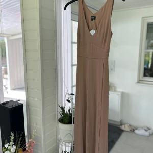 Beige långklänning med cut-out från Board Forum - Säljer en beige långklänning från Board Forum med djup v-ringning och snygg cut-out detalj framtill. Klänningen har breda axelband och är ärmlös, perfekt för sommarens tillfällen. Materialet känns mjukt och följsamt.