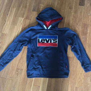 Blå hoodie från Levi's med logga - Säljer en blå hoodie från Levi's med stor broderad logga i blått, vitt och rött framtill. Insidan av huvan är röd och tröjan har känguruficka. Perfekt för dig som gillar klassisk streetstil.