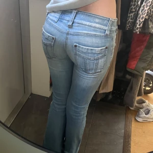 Ljusblå bootcut jeans med slitna detaljer - Säljer ett par ljusblå jeans med bootcut passform och låg midja. Jeansen har slitna detaljer vid bensluten och klassiska bakfickor. Perfekta för en avslappnad och trendig look.