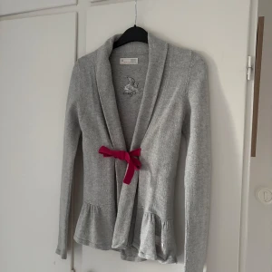 Grå kofta med rosett från Odd Molly - Supersöt grå kofta från Odd Molly, volang nertill och en röd rosett framtill. Stl 0, vilket jag tror motsvarar en XXS-XS. Andvänd men fortfarande bra skick🌸😊