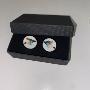 Vita manschettknappar med fiskefluga - Unika manschettknappar med vit bas och motiv av en fiskefluga i blått, svart och brunt. Perfekt accessoar för dig som gillar fiske eller vill ha något annorlunda till skjortan.