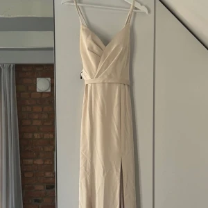 Balklänning champagne light - Elegant, ärmlös klänning med smala axelband och en v-ringning. Klänningen har en figurnära passform med en markering i midjan och en hög slits på ena sidan. Baksidan har en öppen design med korsade band och en dragkedja.  Aldrig använd pga fel storlek!  Färg: Light champagne  Måttbeställd enligt dessa kroppsmått: Byst 87 Under byst 77 Midja 68 Höft 98 Midja till golv 107 Axel till byst 22 Axel till golv 145
