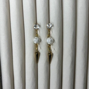 Guldiga örhängen med vita rosor och strass - Säljer ett par unika örhängen med guldiga detaljer, vita rosformade pärlor och glittrande strass upptill. Hängande spetsig detalj längst ner ger en edgy touch. Perfekta för dig som vill sticka ut!