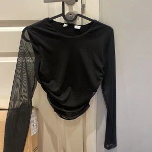 Svart mesh topp med långa ärmar - Säljer en svart mesh topp med långa ärmar. Toppen har en tight passform och är tillverkad i genomskinligt syntetmaterial. Perfekt för att skapa en edgy look.