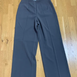 Grå kostymbyxor från NA-KD - Snygga grå kostymbyxor från NA-KD med klassisk rak passform och pressveck. Byxorna har en diskret bakficka och stängs framtill. Perfekta för en stilren look.