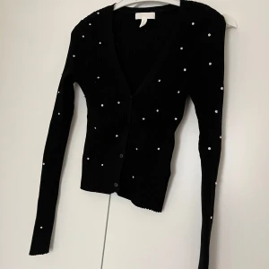 Svart kofta med pärlor - Säljer en svart kofta från H&M med v-ringning och knäppning framtill. Tröjan är dekorerad med små pärlor över hela och har lång ärm. Perfekt för dig som vill ha något stilrent men ändå med en twist.