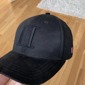 Svart keps från Les Deux - Snygg svart keps från Les Deux med broderad logga framtill och justerbar baksida. Klassisk design med böjd skärm och liten detalj i rött, vitt och blått på sidan. Perfekt accessoar för en stilren look.