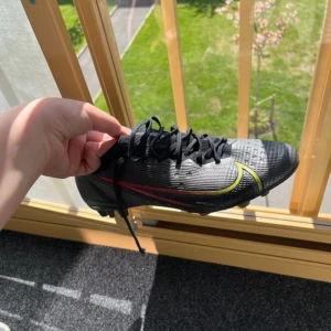 Svarta Nike Mercurial fotbollsskor - Säljer ett par svarta Nike Mercurial fotbollsskor med gula och röda detaljer. Skorna har snörning och är tillverkade i syntetmaterial. Perfekta för dig som vill ha bra grepp på planen och snygg design.