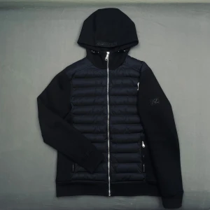 Svart cardigan med huva från AZ - Svart cardigan med huva och dragkedja från AZ. Jackan har quiltade partier framtill och på axlarna, samt släta ärmar. Snyggt broderad logga på ärmen och två fickor med dragkedja. Perfekt för en stilren look.