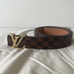 Brunt rutigt bälte från Luviton - 115 cm. Snyggt brunt  bälte från Luvitton. Med rutigt mönster i brunt och mörkbrunt. Helt oanvänd och ren. Perfekt accessoar för att lyfta din outfit. PRISET KAN DISKUTERAS.