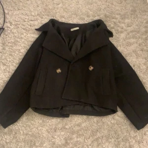 Svart kappa med knappar - Säljer en svart kappa med bred krage och dubbelknäppning framtill. Jackan har två fickor och är något croppad i modellen. Perfekt för dig som vill ha en stilren och klassisk look.