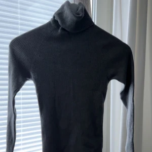 Mörkgrå ribbad polotröja - Säljer en mörkgrå ribbad polotröja med lång ärm. Tröjan är tight i modellen och har en hög polokrage. Perfekt för lager-på-lager och passar till många olika outfits.