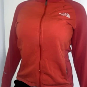 Röd fleecejacka från The North Face - Röd fleecejacka från The North Face med dragkedja framtill och två fickor med dragkedja. Jackan har långärmade ärmar och en sportig passform, perfekt för kyliga dagar.