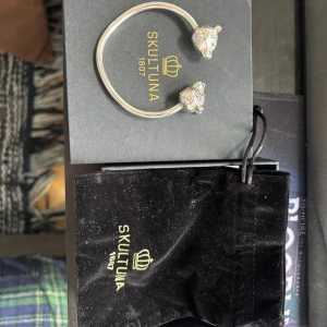 Skultuna silver armband - Unikt armband i silverfärg från Skultuna med två detaljerade björnhuvuden i ändarna. Kommer med originalförpackning och svart tygpåse med Skultuna-logga. Perfekt accessoar för dig som gillar stilrena och djurinspirerade smycken. Aldrig använt!