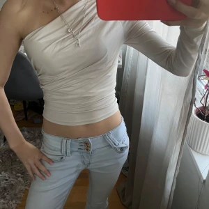 Beige oneshoulder topp - Säljer en beige oneshoulder topp med tight passform. Toppen är croppad och har långa ärmar på ena sidan, vilket ger en trendig och stilren look. Perfekt att matcha med jeans eller kjol.