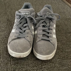 Grå Adidas Campus sneakers - Säljer ett par grå Adidas Campus sneakers med vita ränder och vit häl med svart Adidas-logga. Skorna har snörning och är i mocka med platt sula. Klassisk och stilren modell som passar till det mesta.