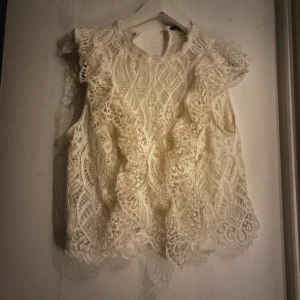Vit spetsblus med volang - Superfin vit blus i spets med volangdetaljer framtill och längs axlarna. Ärmlös modell med rund halsringning och vackert mönstrad spets. Perfekt för att lyfta vilken outfit som helst!