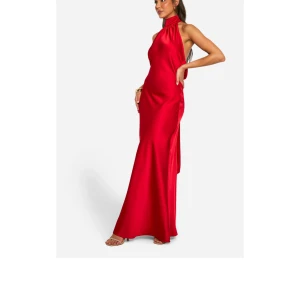 Röd långklänning med halterneck och öppen rygg - Säljer en elegant röd långklänning i satin med halterneck och snygg öppen rygg med banddetaljer. Klänningen har en figurnära passform och en hög slits på sidan. Perfekt för dig som vill sticka ut på festen!
