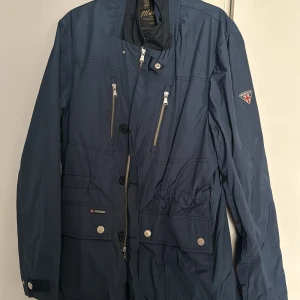 Blå vindjacka från Morris - Snygg blå vindjacka från Morris med dragkedja och knappar framtill. Jackan har flera fickor med lock och dragkedja, samt en patch med Morris-logga på ärmen. Perfekt för blåsiga dagar och stilren look. Nypris på dessa jackor är 3000kr. Jackan har alldrig blivit använt ute då jag inte haft tid att använda just den hära jackan.