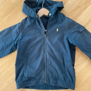 Blå vindjacka från Polo Ralph Lauren - Snygg blå vindjacka från Polo Ralph Lauren med huva och dragkedja framtill. Jackan har två fickor fram och en ärmficka med knapp. Klassisk logga broderad på bröstet. Perfekt för blåsiga dagar.
