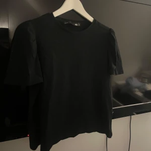 Svart topp med puffärm från 157 - En svart topp från 157 med rund halsringning och korta puffärmar. Enkel och stilren design som passar till många olika outfits. Tillverkad i mjukt material för en bekväm känsla.