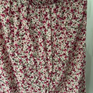 Blommig långkjol med knappar - Säljer en långkjol med blommigt mönster i rosa, rött och grönt på vit botten. Kjolen har knappar framtill och ett luftigt, flowy tyg som passar perfekt för varmare dagar.