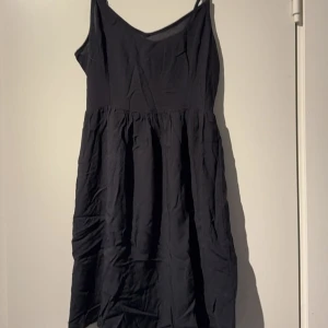 Svart axelbandsklänning - En svart klänning med smala axelband och v-ringad halsringning. Klänningen har en luftig passform med rynkad midja och är tillverkad i ett lätt tyg, perfekt för varma dagar.