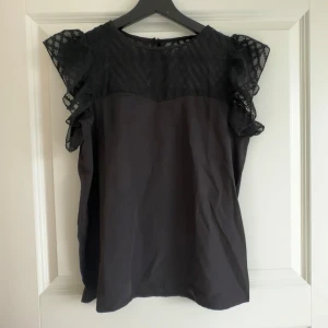 Svart blus med spets och volang - Säljer en svart blus med spets upptill och volangdetaljer på axlarna. Blusen är ärmlös och har en rund halsringning. Perfekt för dig som gillar feminina detaljer och stilren design.