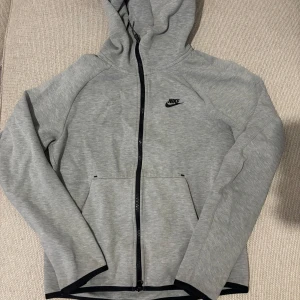Grå hoodiejacka från Nike - Säljer en grå hoodiejacka från Nike med svart dragkedja och logga på bröstet. Jackan har huva, långa ärmar och två fickor framtill. Perfekt för en sportig och avslappnad stil.