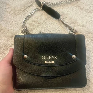 Svart axelväska från Guess - Säljer en stilren svart axelväska från Guess med silverfärgad kedja. Den är lite trasig i utkanten, men inget som inte går att fixa.