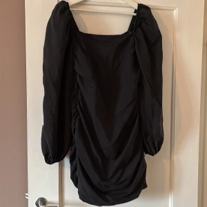 Svart puffärmad klänning från Nelly - Säljer en svart kort klänning från Nelly med puffiga långa ärmar och rynkade detaljer framtill. Klänningen har fyrkantig urringning och dragkedja i ryggen. Perfekt för dig som vill sticka ut med en trendig look.
