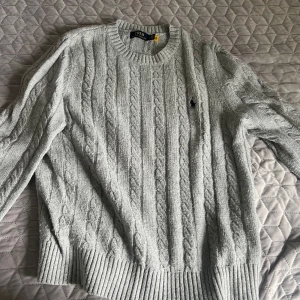 Ljusgrå kabelstickad tröja från Polo Ralph Lauren - Säljer en klassisk ljusgrå kabelstickad tröja från Polo Ralph Lauren med rund halsringning och broderad logga på bröstet. Tröjan har långa ärmar och breda ribbade muddar vid ärmslut och nederkant.